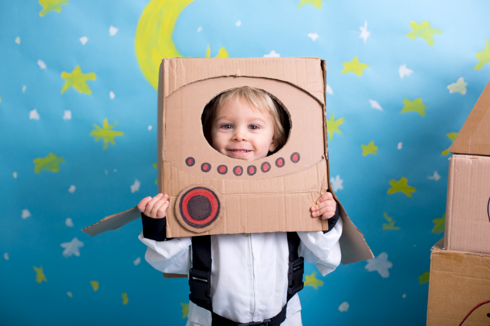 Sweet toddler boy, dressed as an astronaut, playing at home with cardboard rocket and handmade helmet from box