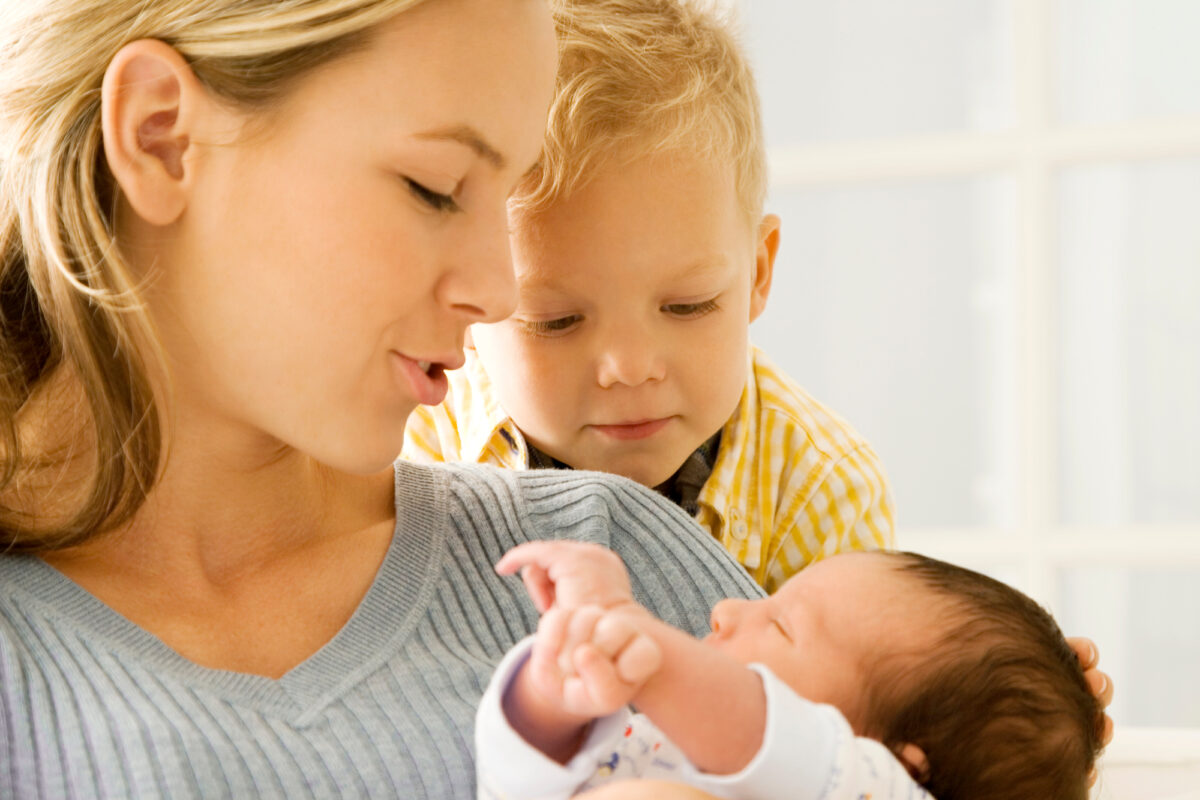 mom holding newborn with 2 year old toddler looking over shoulder