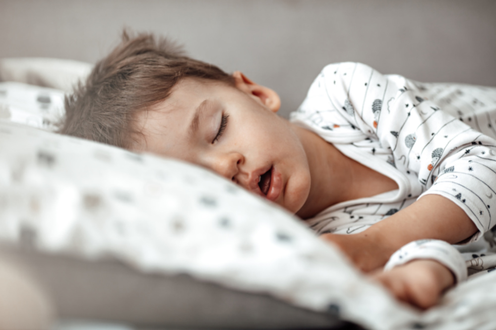 Little blond boy sleeping in his bed. Restless sleep of a baby with a cold. The kid sleeps with his mouth open. My little boy sleeping by the light of the window.