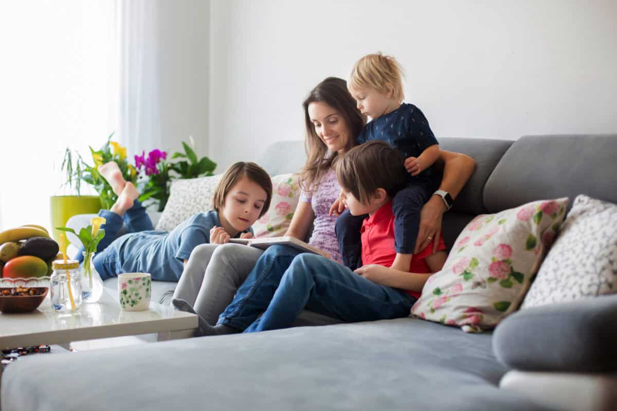 Young woman, mother with three kids, reading a book at home, hugging and laughing, family happiness